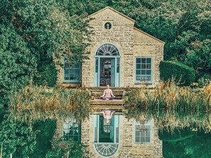 A stone building with a central arched window and a person meditating on the steps. - West Lexham Wellness and Retreat Centre (King's Lynn)
