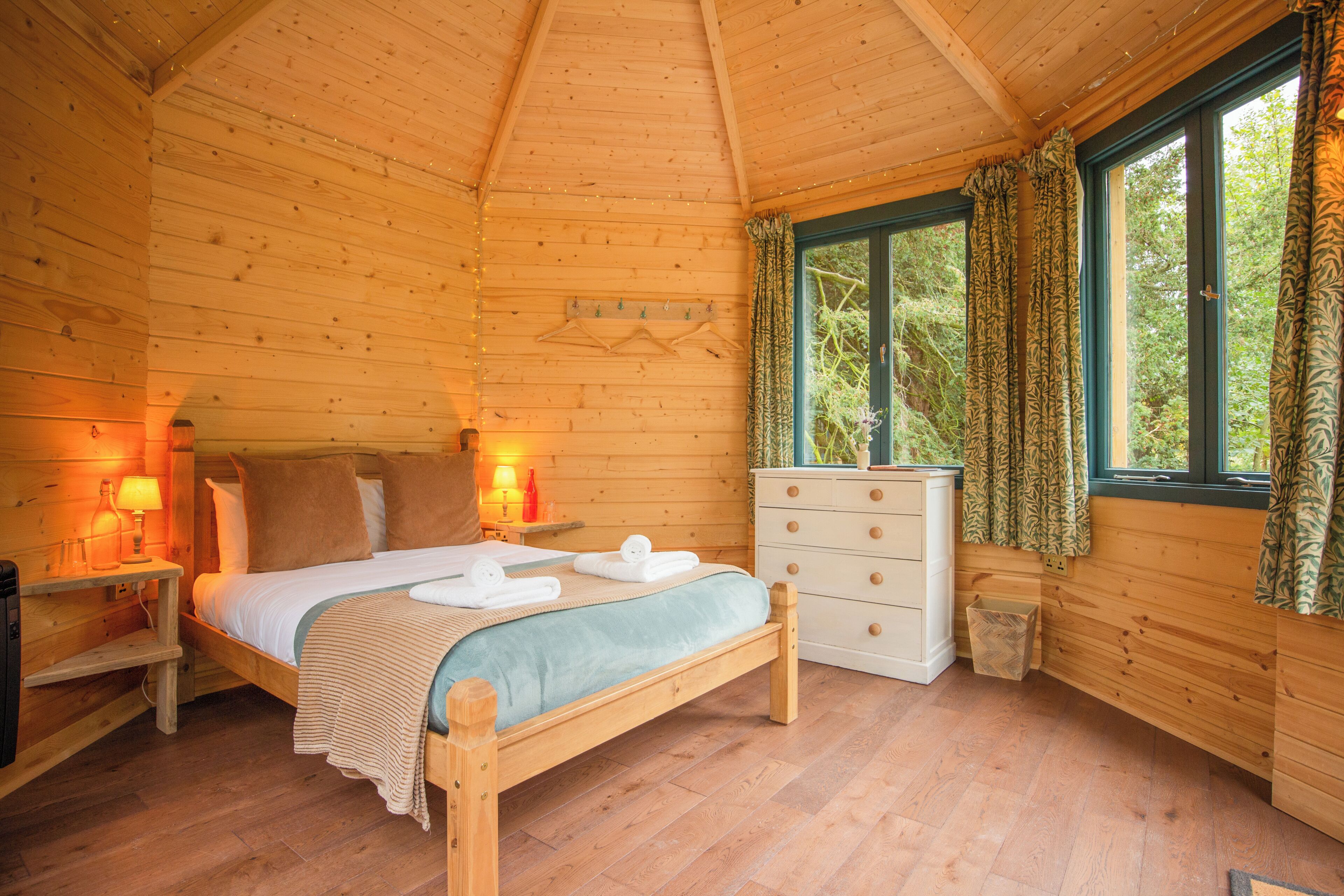 A wooden cabin bedroom with a bed, bedside lamps, a dresser, and a window with curtains.