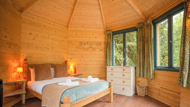 A wooden cabin bedroom with a bed, bedside lamps, a dresser, and a window with curtains.