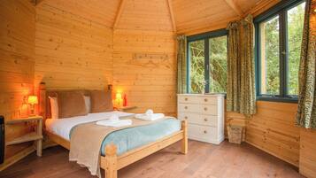 A wooden cabin bedroom with a bed, bedside lamps, a dresser, and a window with curtains.