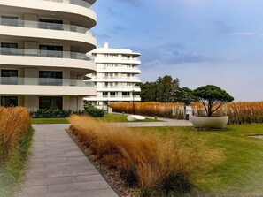 A modern multi-story residential building with a curved facade, a landscaped garden with tall grasses, and a large planter with a tree. - Apartment in Rogowo Near Beach and Lake (Trzebiatow)