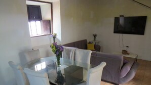 Uma área de refeições com mesa de vidro e cadeiras brancas, um sofá e uma TV de tela plana fixada na parede.