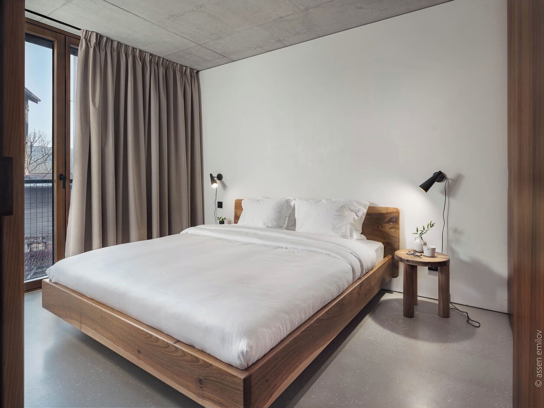 Un dormitorio moderno con una cama de madera, dos mesitas de noche con lámparas, vistas a través de un ventanal grande y un techo de hormigón.