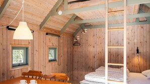 Une cabane en bois comprenant un coin repas, deux fenêtres, un lit et un luminaire suspendu.