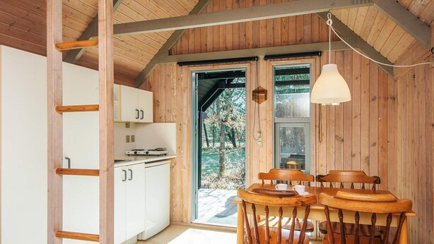 L’intérieur chaleureux d’un chalet avec un coin repas en bois, une cuisine dotée de meubles blancs et une grande fenêtre offrant une vue sur l’extérieur.
