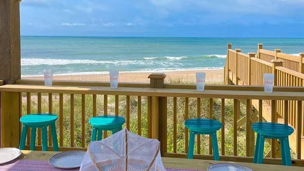 Una terraza de madera con taburetes turquesa y una mesa, con vista a una playa que tiene una valla de madera y un cielo despejado.