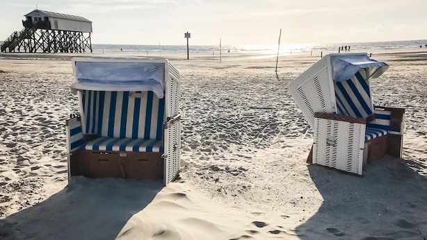 Two beach chairs with striped cushions on a sandy beach, a pier structure in the background, and a clear sky. - Ferienwohnung in St. Peter-ording (St. Peter-Ording)