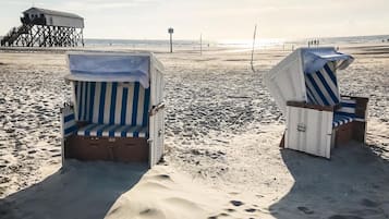 Dos sillas de playa con cojines a rayas sobre una playa arenosa, con una estructura de muelle al fondo y un cielo despejado.