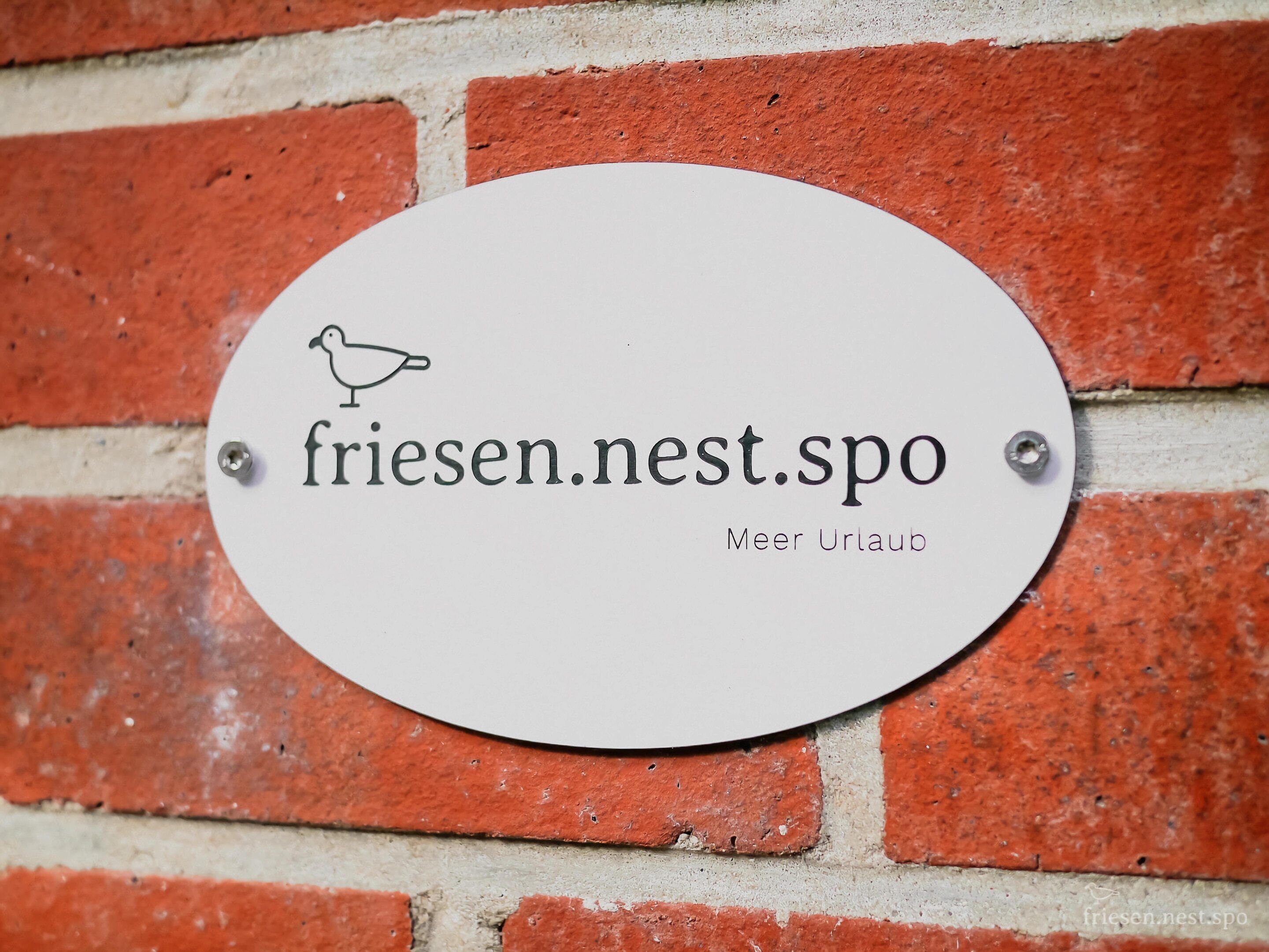 A white sign with the text "friesen.nest.spo" and a bird icon, mounted on a red brick wall.