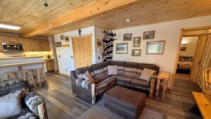 A cozy living room with a sectional sofa, a wooden coffee table, and a patterned rug. There is a kitchen area with wooden cabinets and a microwave. The room has wooden flooring and a ceiling with exposed beams. - OPEN YEAR ROUND - 3 Bedroom Lake San Cristobal Condo on the Alpine Loop (Lake City)