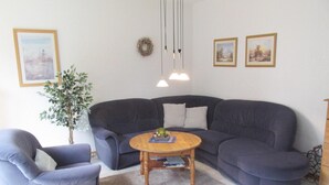 A living room with a dark blue sectional sofa, a round wooden coffee table, and a grey armchair. There are two wall-mounted pendant lights, a green plant, and framed pictures on the wall. - Ferienunterkunft in Hagermarsch (Hagermarsch)
