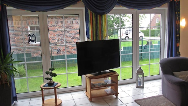 A living room with a TV on a wooden stand, a small side table, a grey sofa, and a large window with curtains. - Ferienunterkunft in Hagermarsch (Hagermarsch)