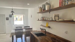 Een eethoek met een houten tafel en stoelen, een keukenblad met een spoelbak, en bovenliggende planken met keukengerei en decoratieve objecten.