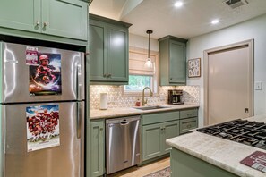 Une cuisine équipée d’appareils électroménagers en acier inoxydable, de meubles de rangement verts, d’un lave-vaisselle et d’une fenêtre avec des stores.
