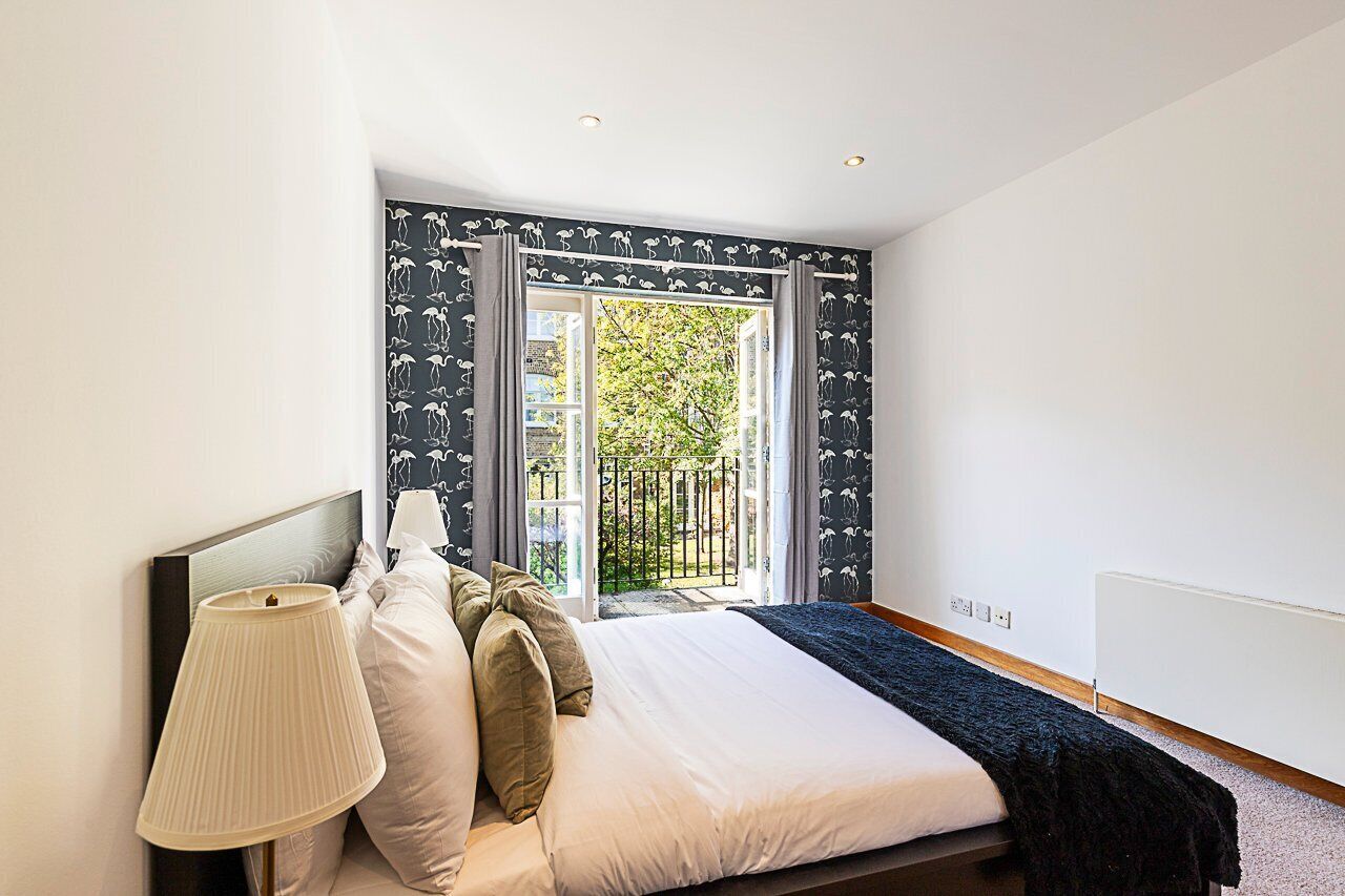 A bedroom with a bed, a nightstand, a lamp, a window with curtains, and a view of greenery outside.