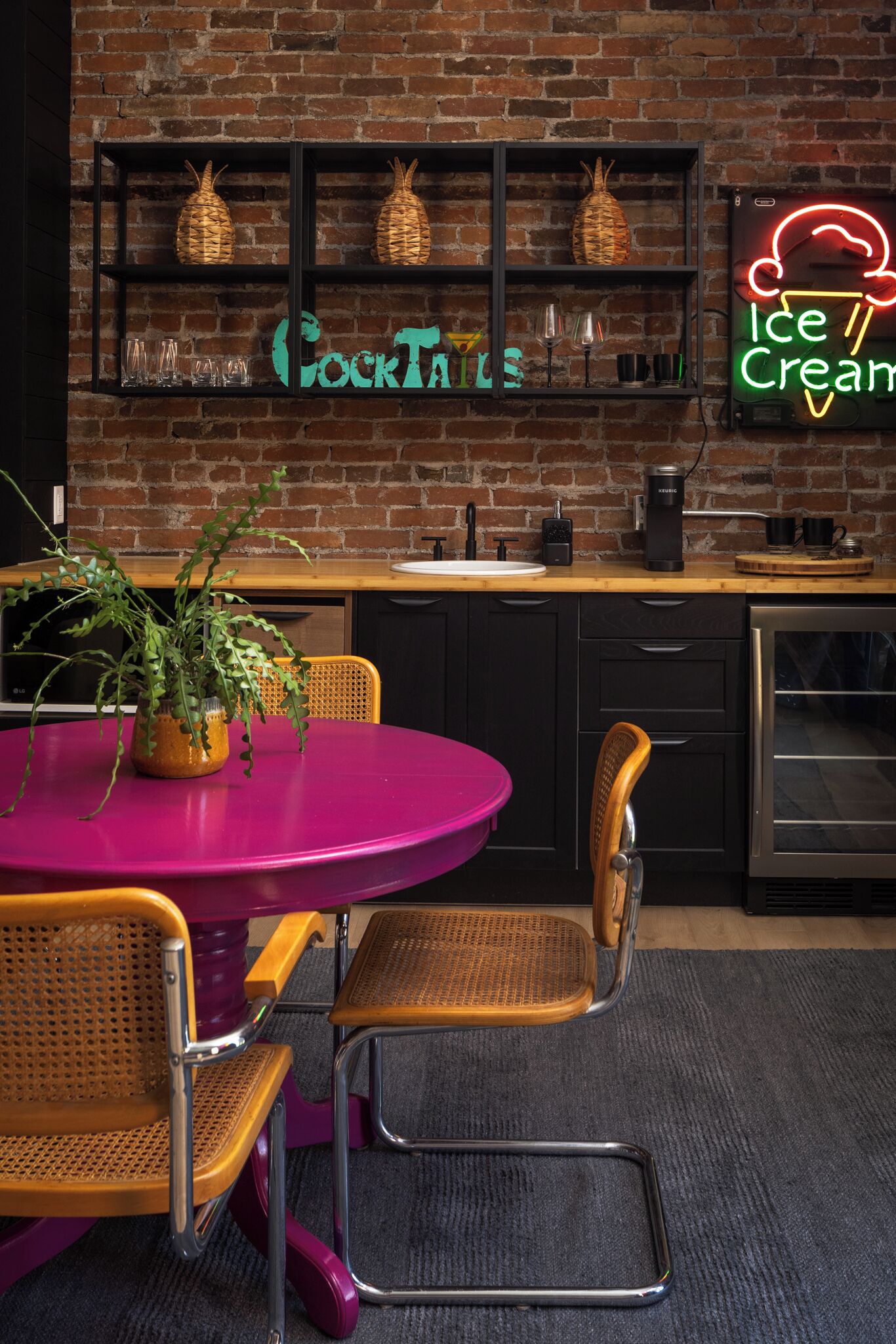 A kitchen with a brick wall, a neon sign, a wooden shelf with pineapples, and a pink table with chairs.