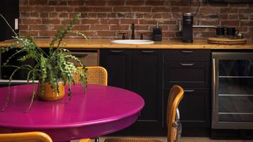 A kitchen with a brick wall, a neon sign, a wooden shelf with pineapples, and a pink table with chairs.