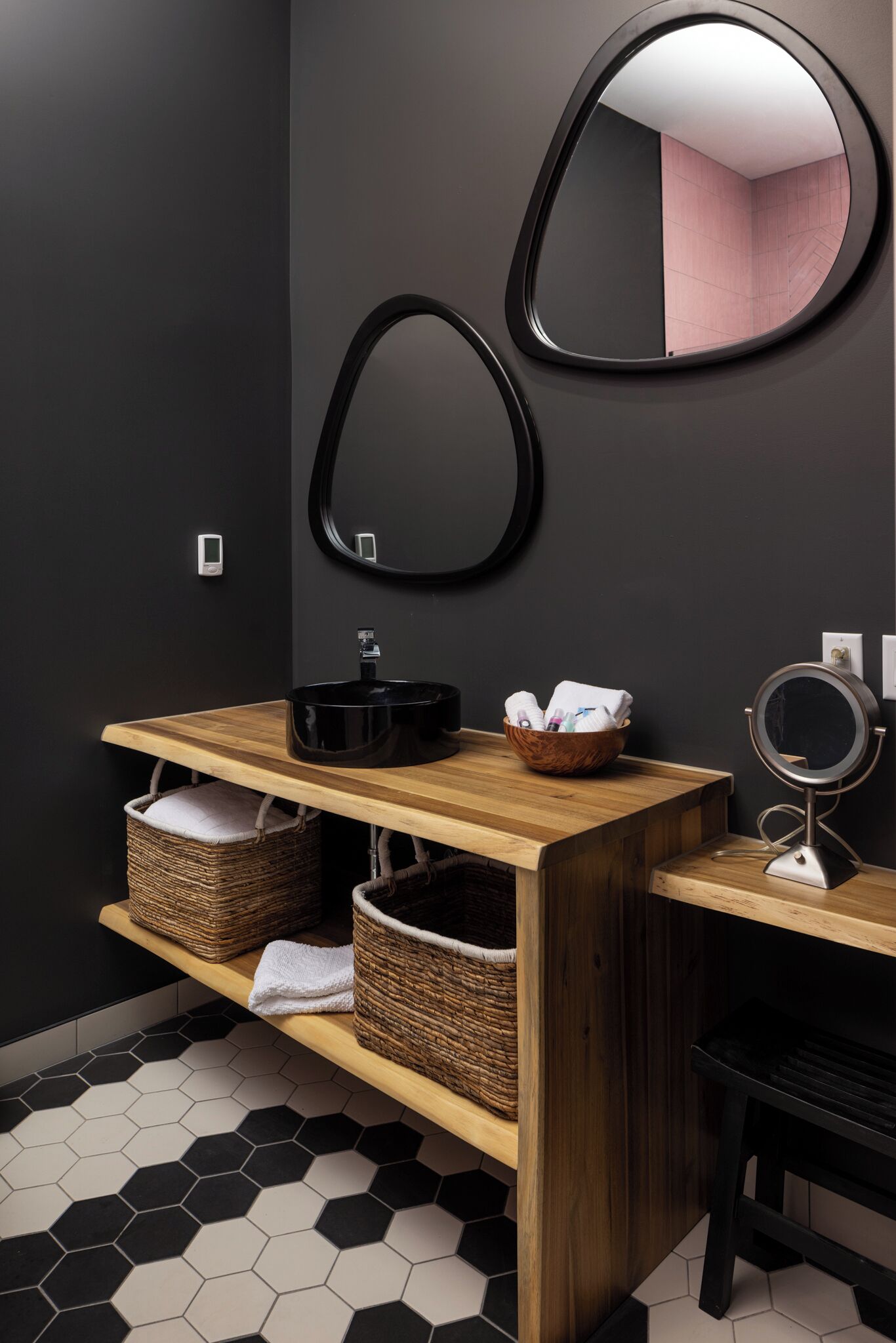 A modern bathroom with a wooden vanity, black and white hexagonal tiles, a round mirror, and two wicker baskets.