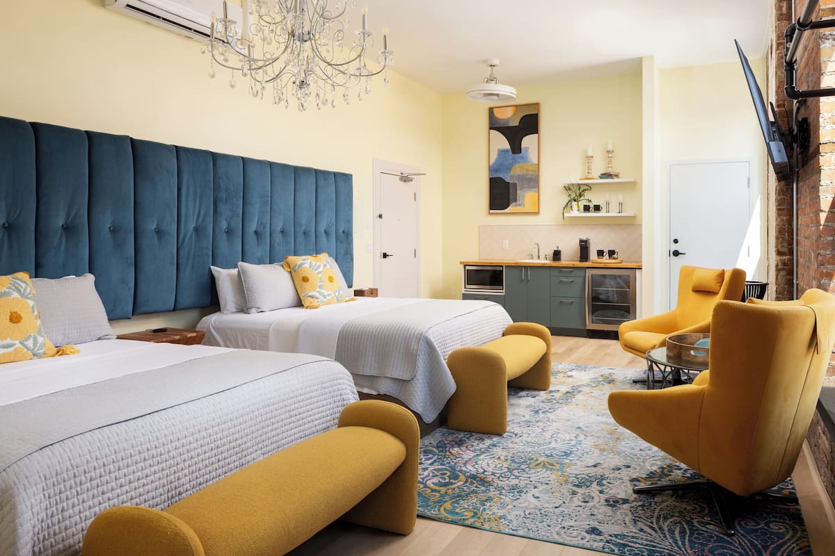 A modern hotel room with two beds, a sitting area with a yellow chair, a chandelier, and a kitchen area.