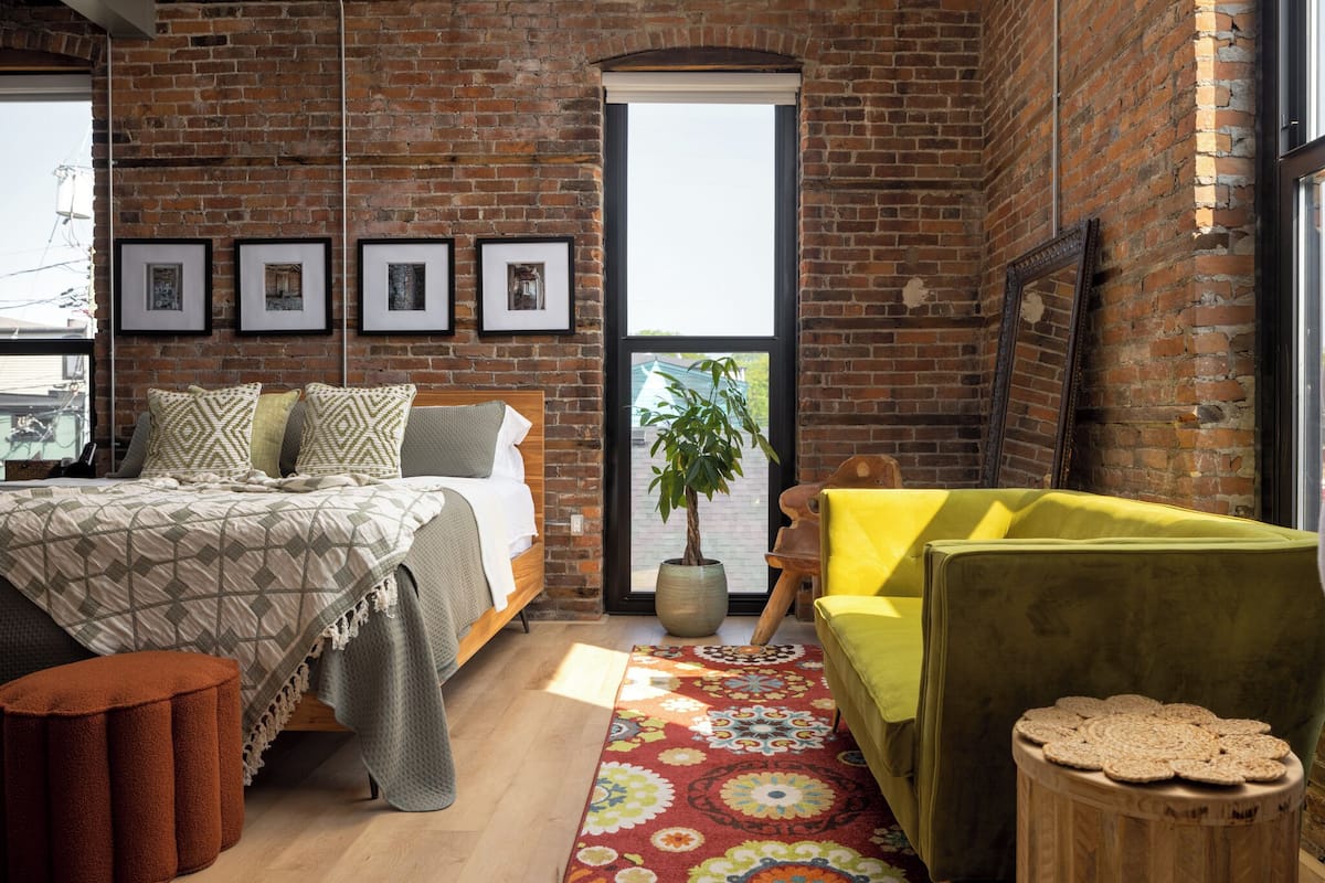 A room with a brick wall, a bed with patterned bedding, a yellow armchair, a wooden side table, and a large window with a view.