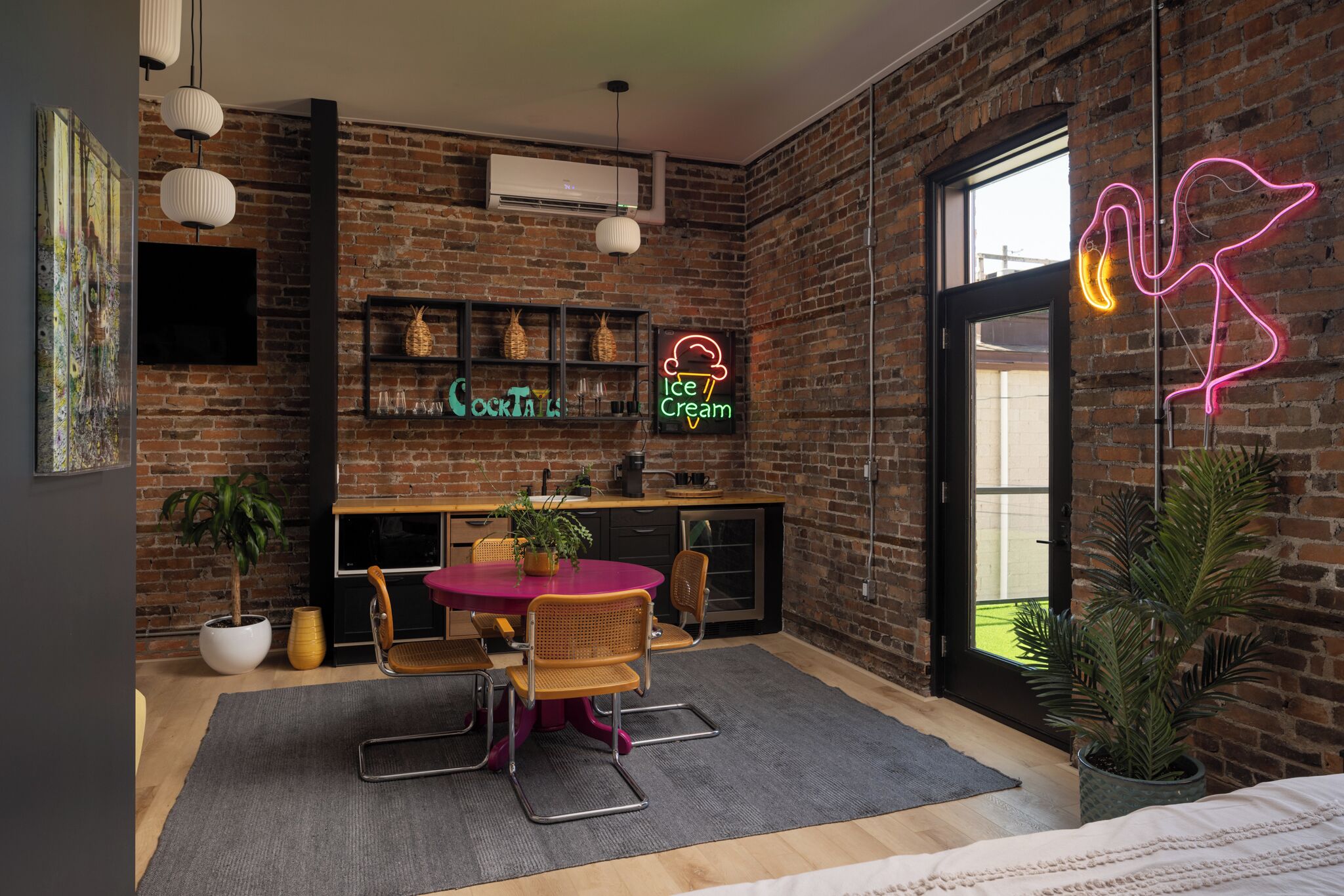 Una cocina moderna con pared de ladrillo, un letrero de neón que anuncia cócteles y helados, una mesa redonda con sillas y un ventanal grande.