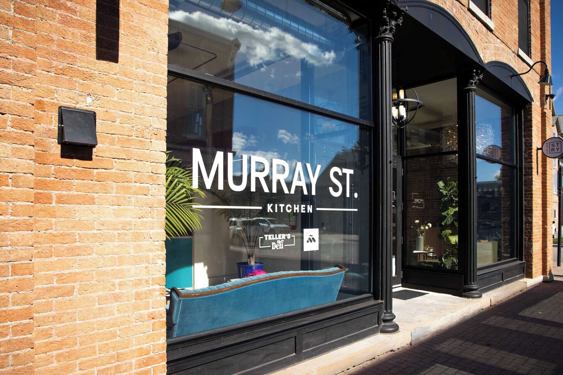 Tiilestä rakennettu rakennus, jossa on suuri ikkuna, ja ikkunassa näkyy teksti: ”MURRAY ST. KITCHEN” ja ”TELLER’S DELI”.