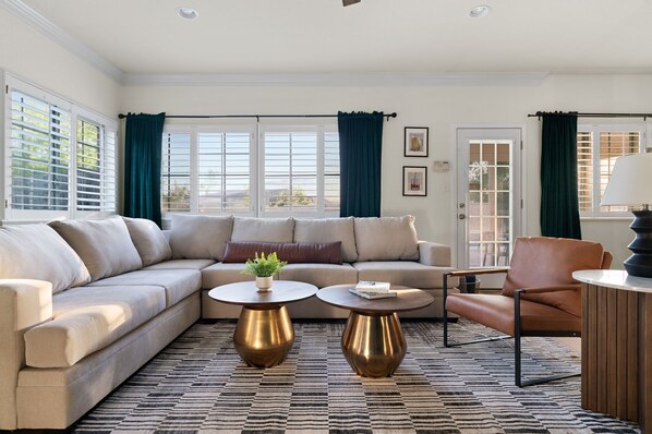 A modern living room with a beige sectional sofa, two round coffee tables, a leather armchair, and a door with a view. - The Mad Hatter Heated Pool Gameroom  (Pflugerville)