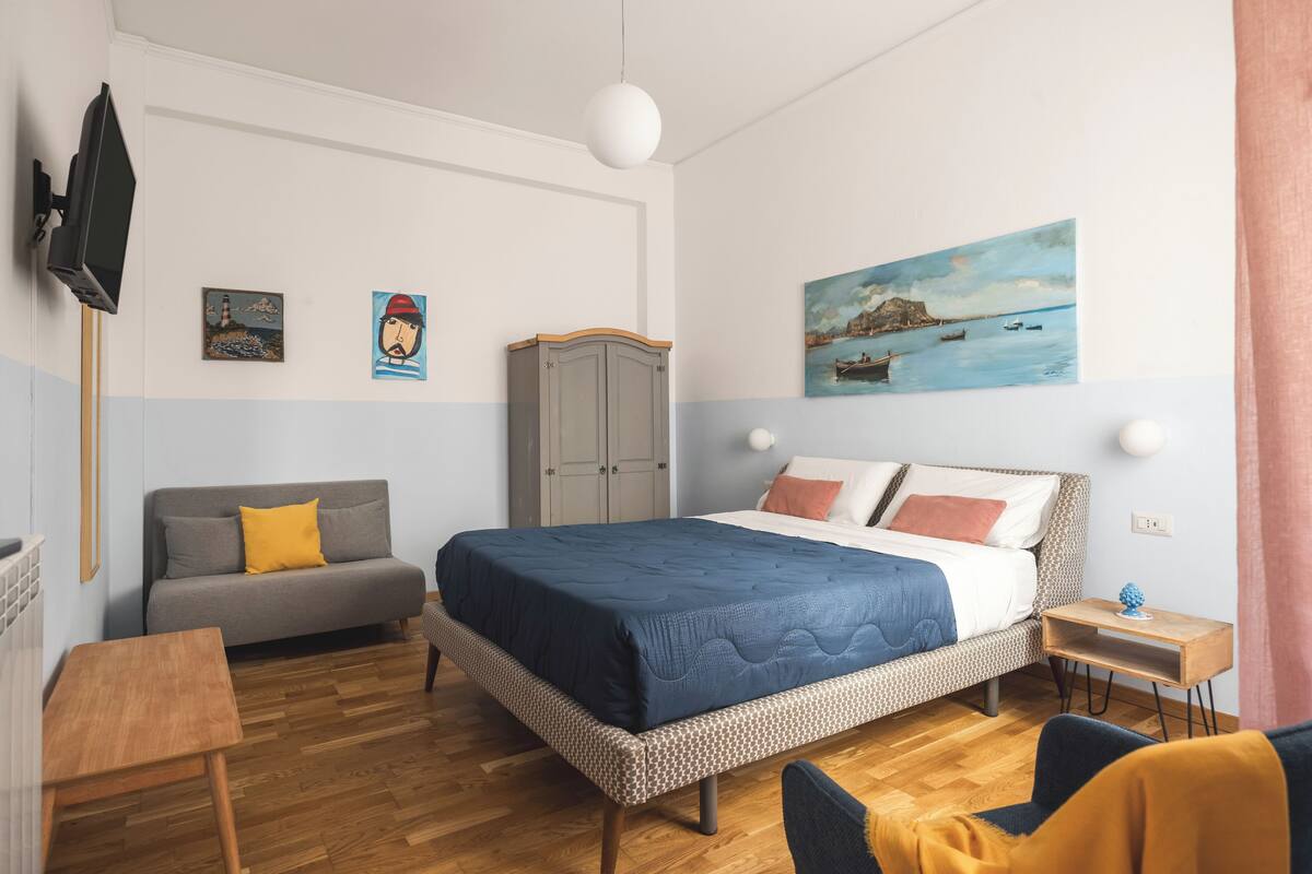 A bedroom with a bed, a grey sofa, a wooden table, and a TV on the wall.