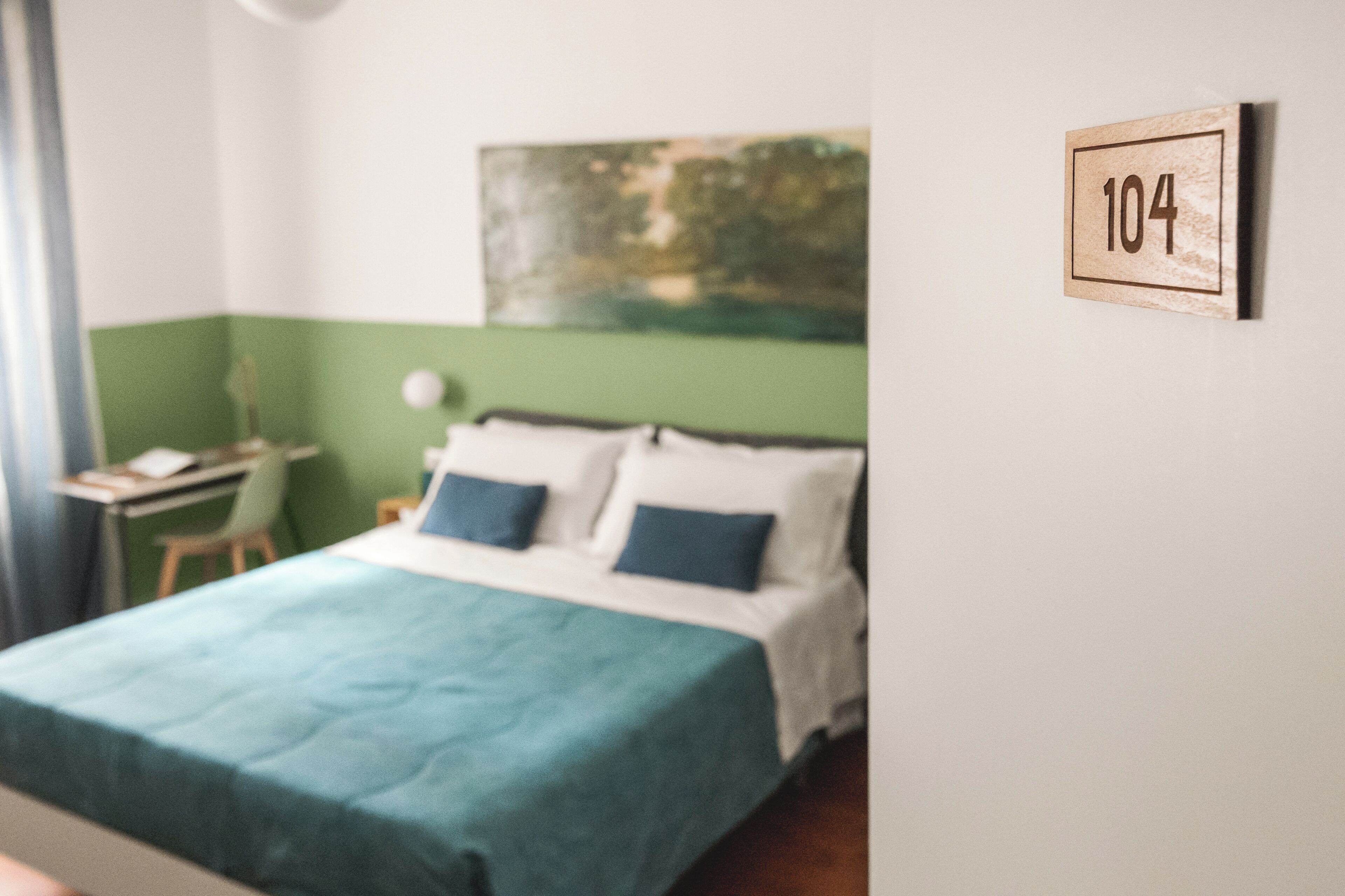 A bedroom with a bed, a desk, and a chair. There is a painting on the wall and a number sign.