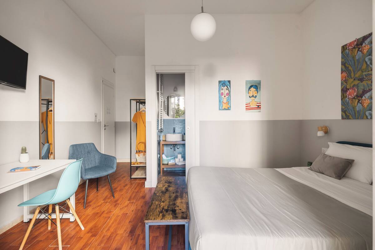 A modern bedroom with a bed, a desk, and a chair. There is a mirror, artwork on the wall, and a television.