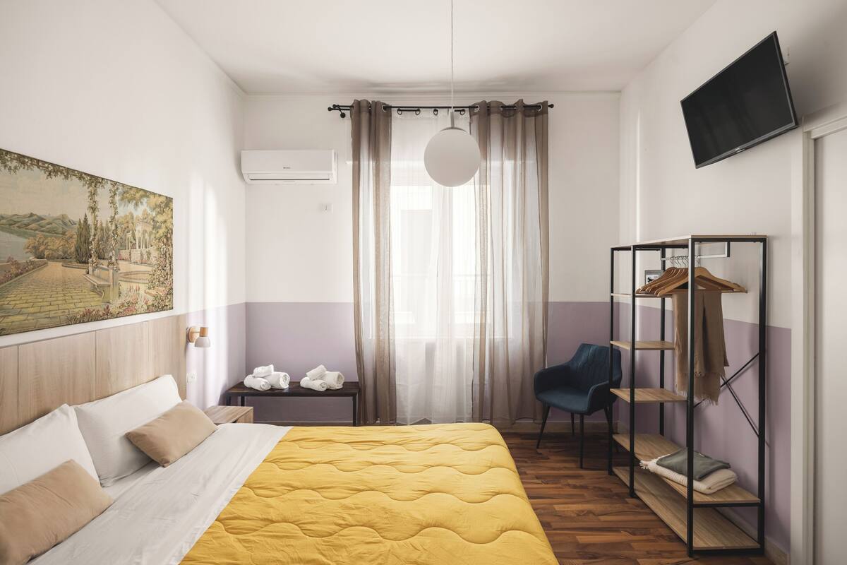 A hotel room with a bed, a desk, a chair, a wardrobe, and a painting on the wall.