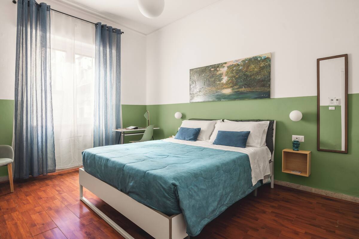 A bedroom with a bed, a desk, a chair, a painting, and a nightstand.