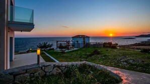 Vista del atardecer desde un balcón con vista al mar, con un pequeño pabellón y un edificio con fachada de vidrio.
