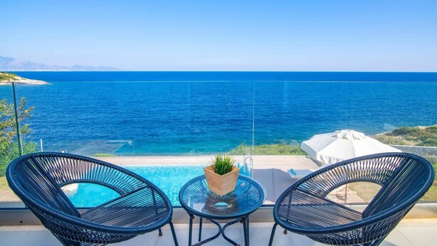 Un balcón con dos sillas de mimbre, una mesita y una planta en maceta, con vistas a un mar azul cristalino y una costa lejana.