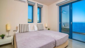 Un dormitorio con cama, dos ventanas, una mesita de noche con lámpara y vista al mar a través de un ventanal.