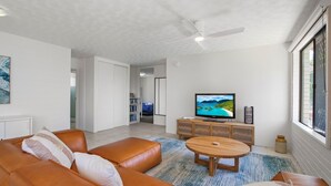 A modern living room with a leather sofa, a wooden coffee table, and a flat-screen TV on a wooden stand. There is a bookshelf and a dining area visible in the background.