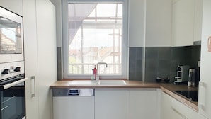 A modern kitchen with white cabinetry, a built-in oven, a dishwasher, and a window with blinds. - MoMa Escape Kecskemét (Kecskemét)