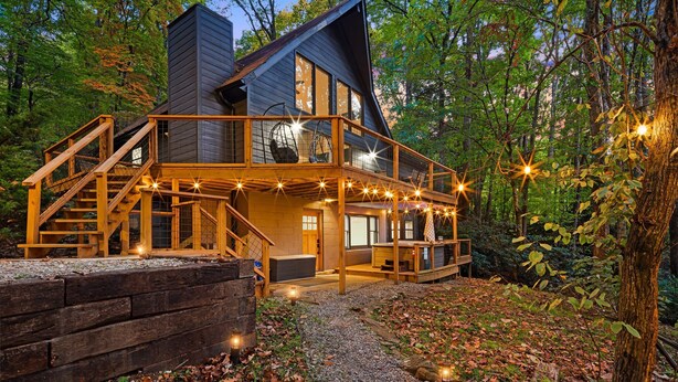 A two-story log cabin with a balcony and string lights, surrounded by a forest. - Summer Special: Hot Tub, Game Room, Cold Plunge & Coffee Bar! (Maggie Valley)