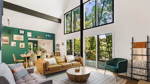 A spacious living room with a high ceiling, a large window overlooking a deck, a green wall with framed pictures, and a modern seating arrangement. - Summer Special: Hot Tub, Game Room, Cold Plunge & Coffee Bar! (Maggie Valley)