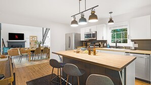 A modern kitchen with a central island, bar stools, a dining area with a sofa, and a TV mounted on the wall. - Summer Special: Hot Tub, Game Room, Cold Plunge & Coffee Bar! (Maggie Valley)
