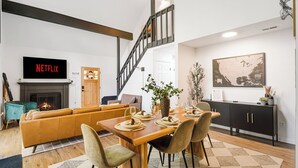 A modern living room with a fireplace, a dining table set for six, and a staircase leading to another level. - Summer Special: Hot Tub, Game Room, Cold Plunge & Coffee Bar! (Maggie Valley)