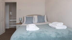 A neatly made bed with white linens and a light blue bedspread, a bedside table with a lamp, and a door leading to a hallway. - Cosy Flat in Leigh | Sea View - Pass the Keys (Southend-on-Sea)