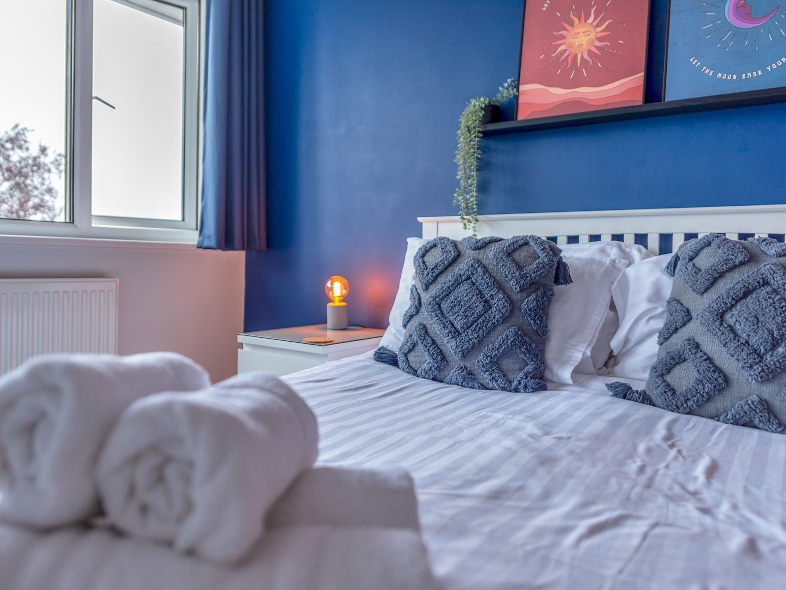 A neatly made bed with rolled white towels, a bedside lamp, and a wall with a blue wall and a window.