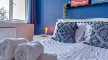 A neatly made bed with rolled white towels, a bedside lamp, and a wall with a blue wall and a window.