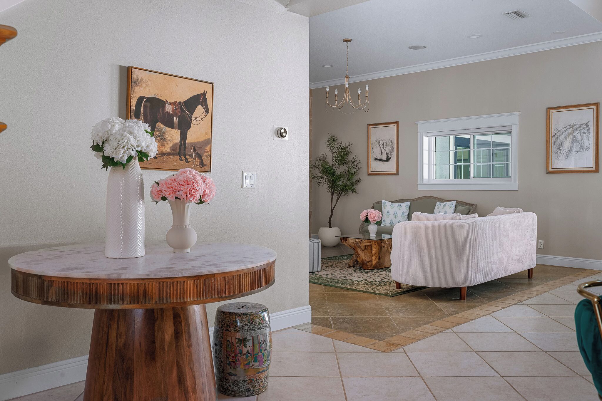 A room with a round wooden table, a white vase with flowers, a bed with a white cover, and a window with a view.