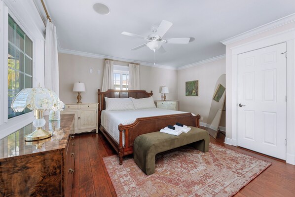 A bedroom with a large bed, a ceiling fan, a window with curtains, a bedside table with lamps, and a door.