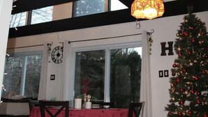 Un comedor con mantel rojo, sillas de madera, un árbol de Navidad y un amplio ventanal con vigas negras.