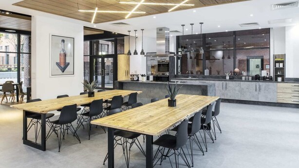 A modern dining area with wooden tables and black chairs, a kitchen with stainless steel appliances, and a large window providing natural light. - Modern Apartments in Hollis Croft Sheffield (Sheffield)