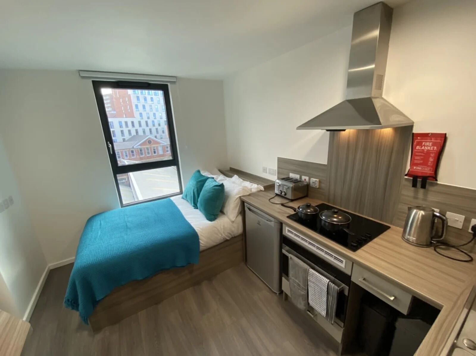 A compact hotel room with a bed, a small kitchenette, and a window with a city view.