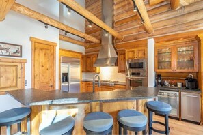 A kitchen with wooden cabinets, a central island with stools, stainless steel appliances, and a high ceiling with exposed beams.